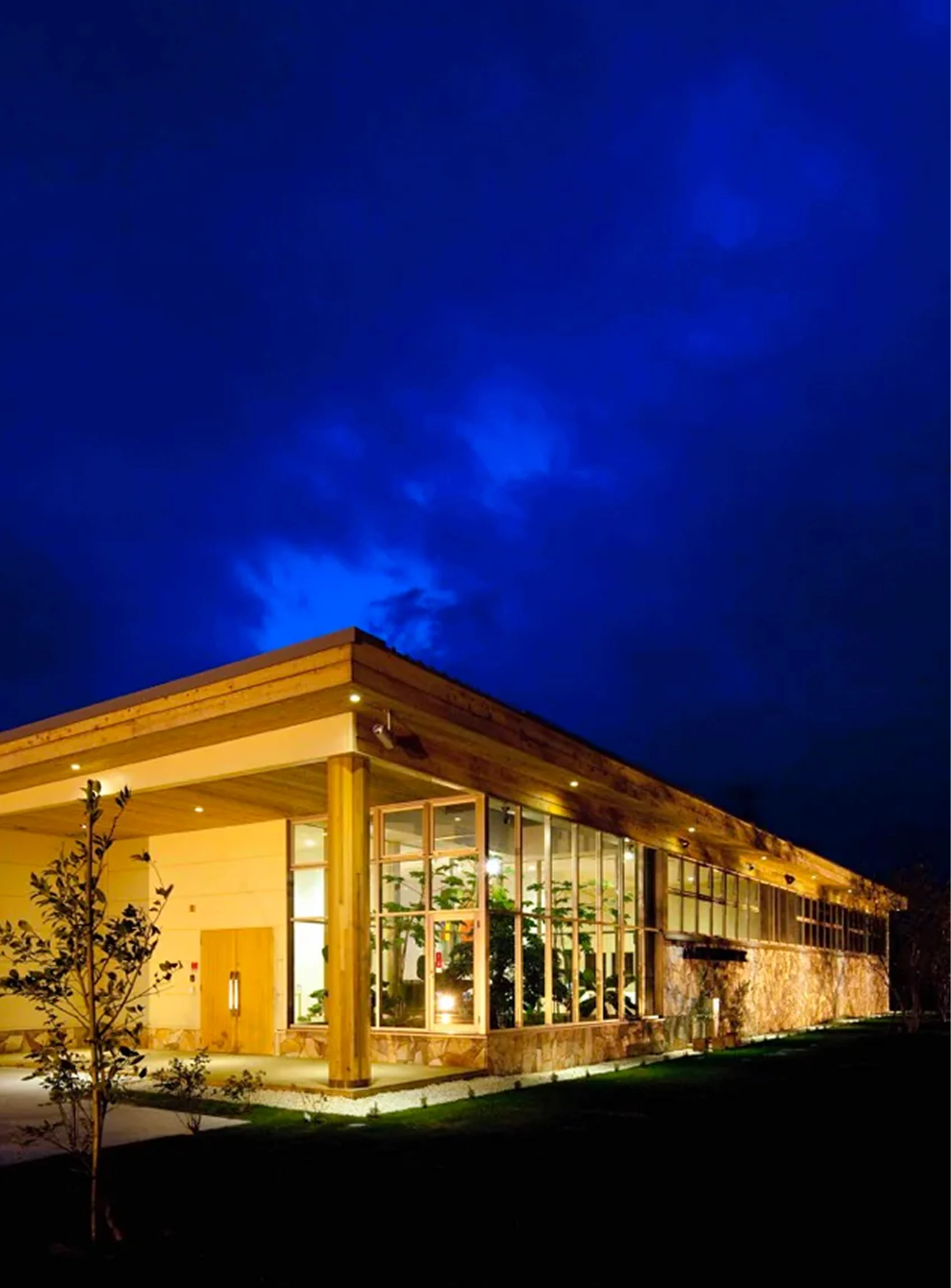 A modern, single-story building with large windows, illuminated at night against a deep blue sky.