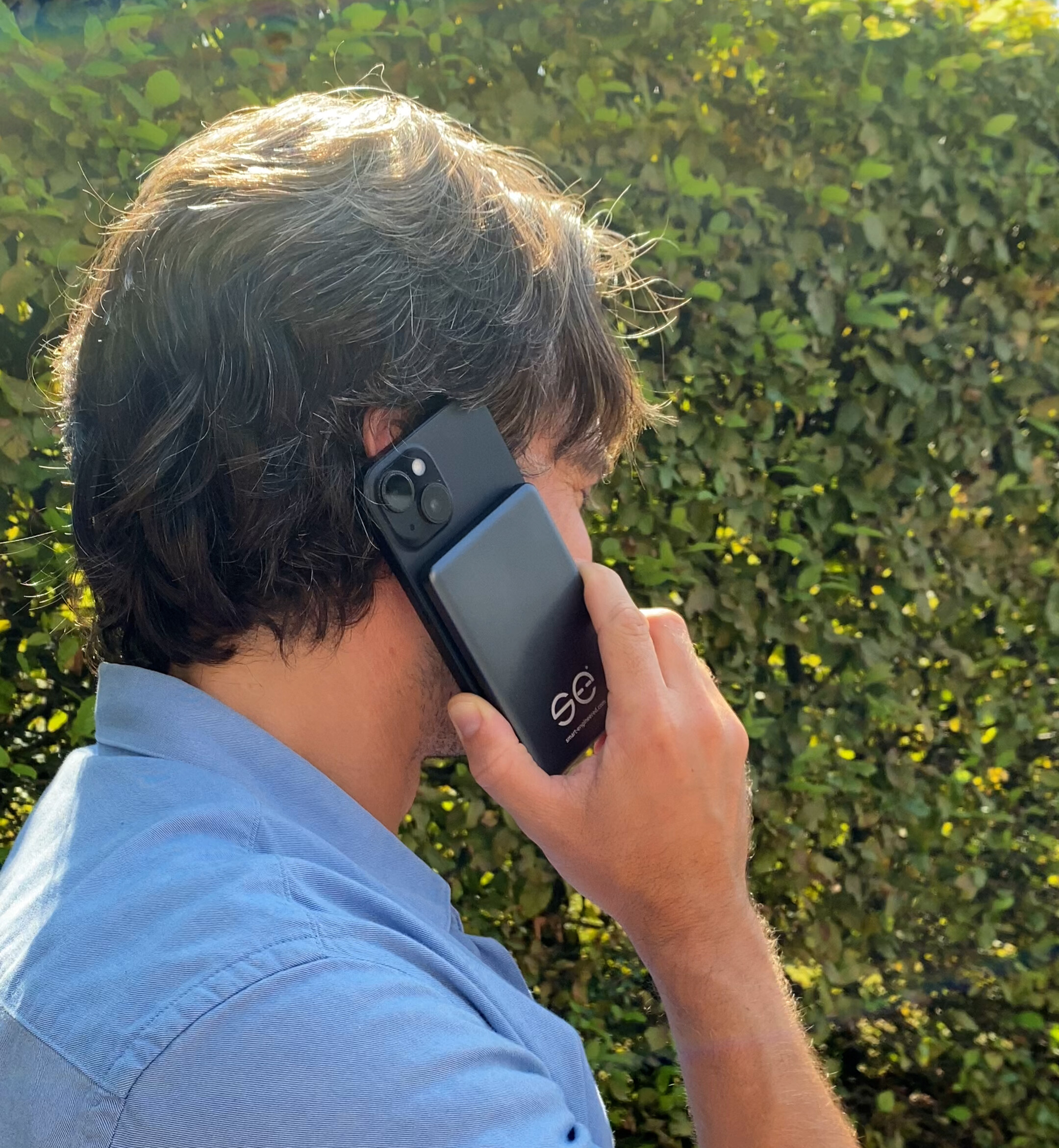 Person holding a smartphone to their ear, standing in front of a leafy hedge.