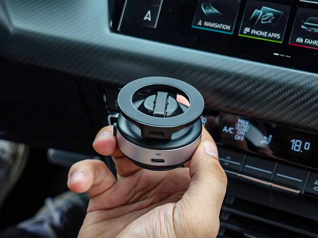 Hand holding a small round device inside a car, near the dashboard.