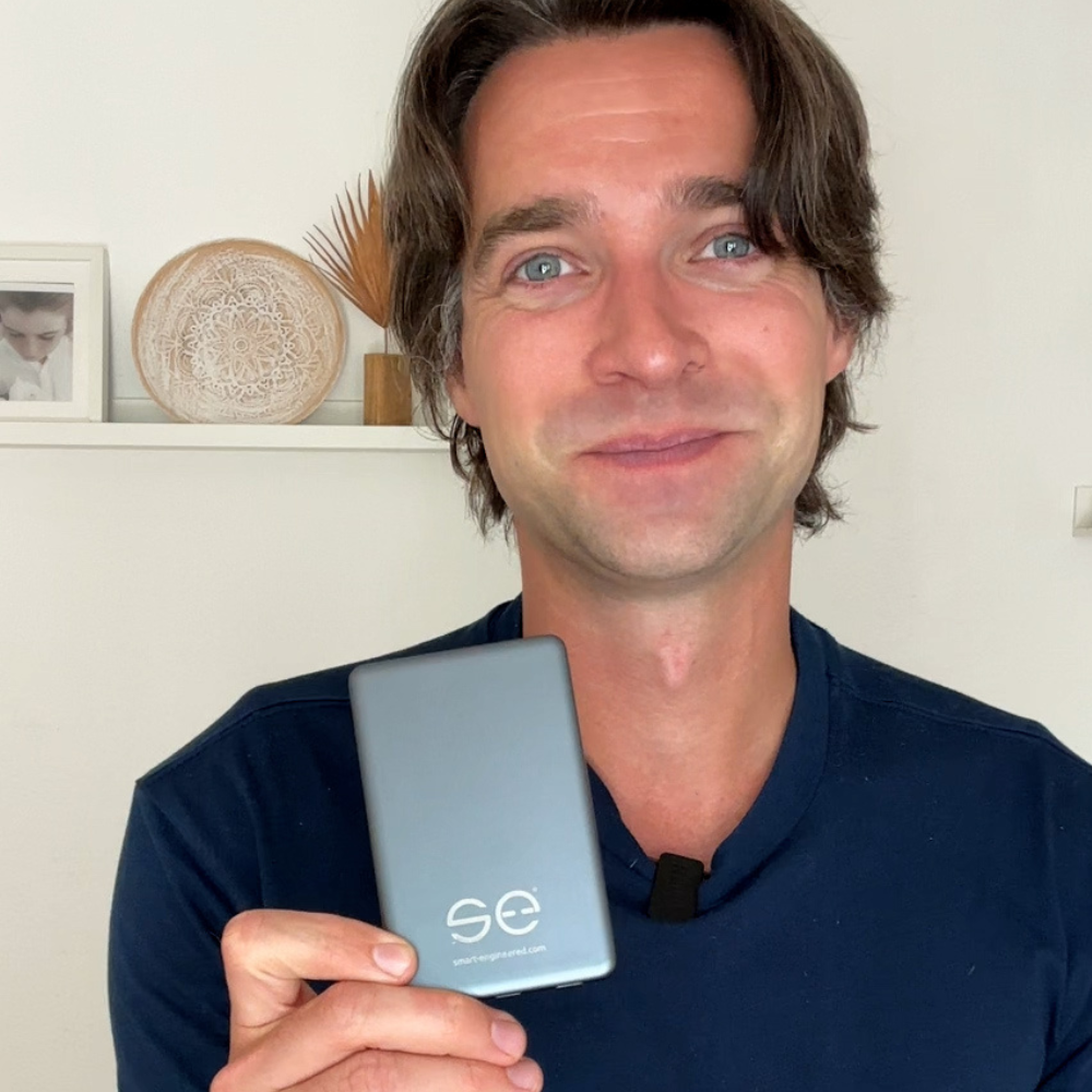 Person holding a small device with 'SE' logo, standing in front of a shelf with decor.