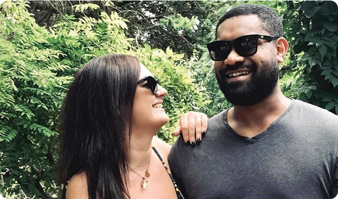 A smiling couple wearing sunglasses outdoors, surrounded by greenery.