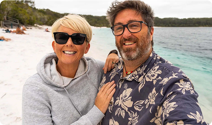 A couple smiling on a beach, with trees and water in the background.