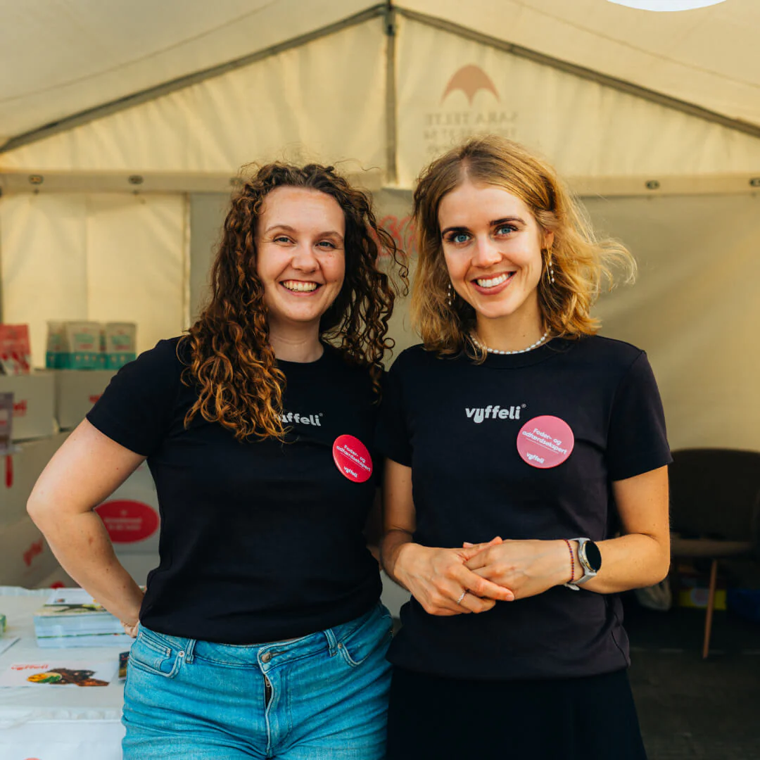 Two women wearing matching shirts stand smiling in a tent with books and boxes visible.