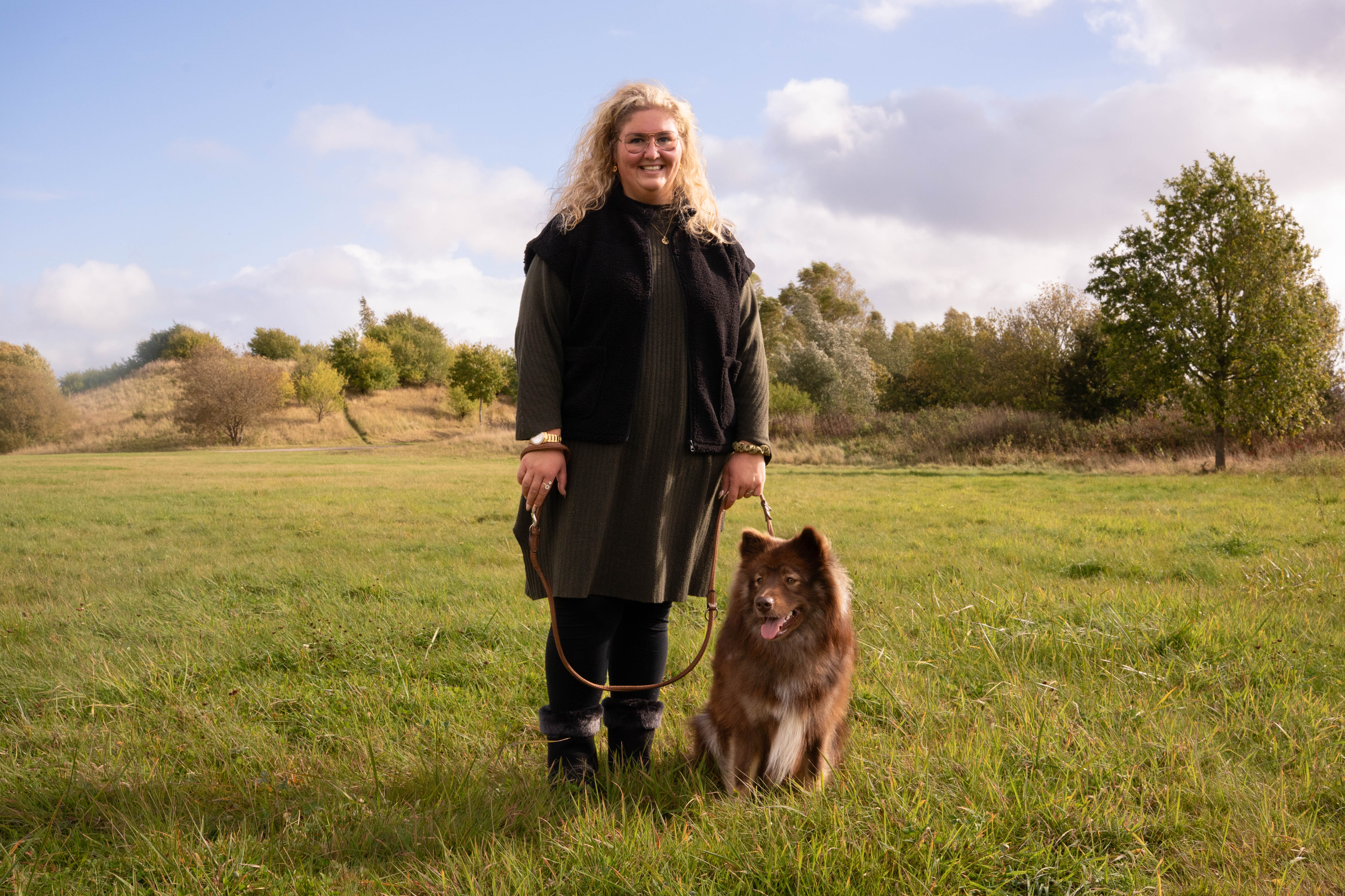 Person standing with a brown dog on a grassy field, trees in the background.