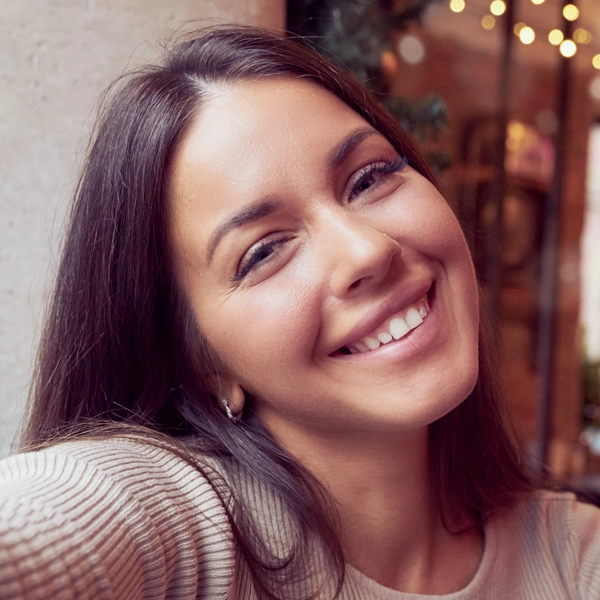 Smiling person taking a selfie indoors with blurred lights in the background.