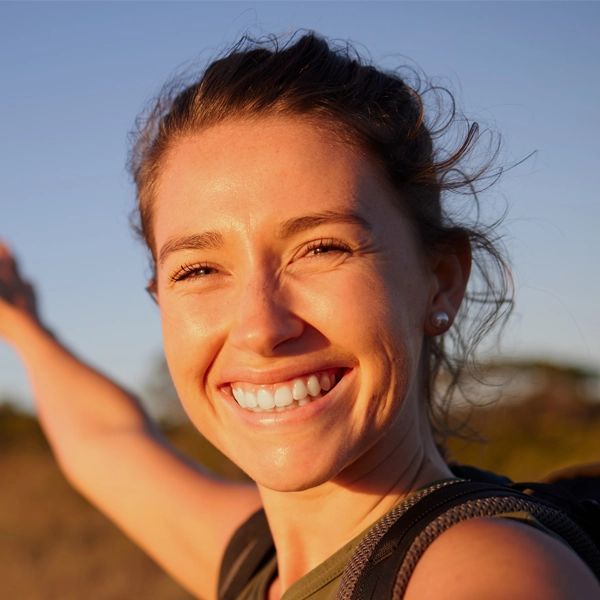 Smiling person outdoors with a backpack, arm outstretched, in warm sunlight.