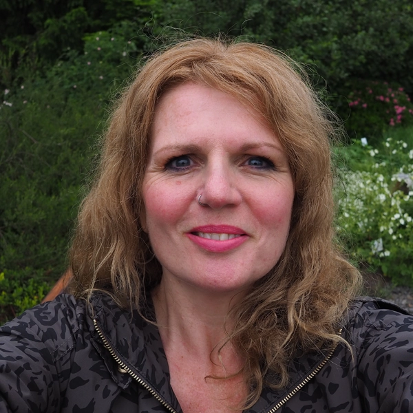 Person with curly hair smiling outdoors with greenery in the background.