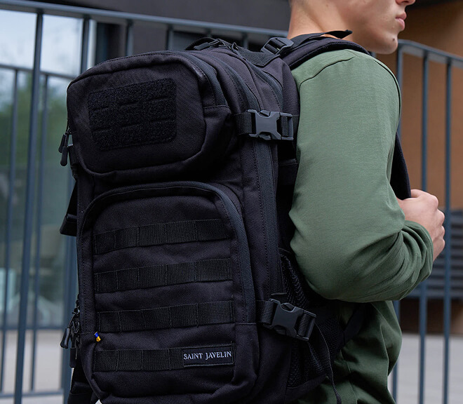 Person wearing a black tactical backpack with a green shirt, in an outdoor setting.