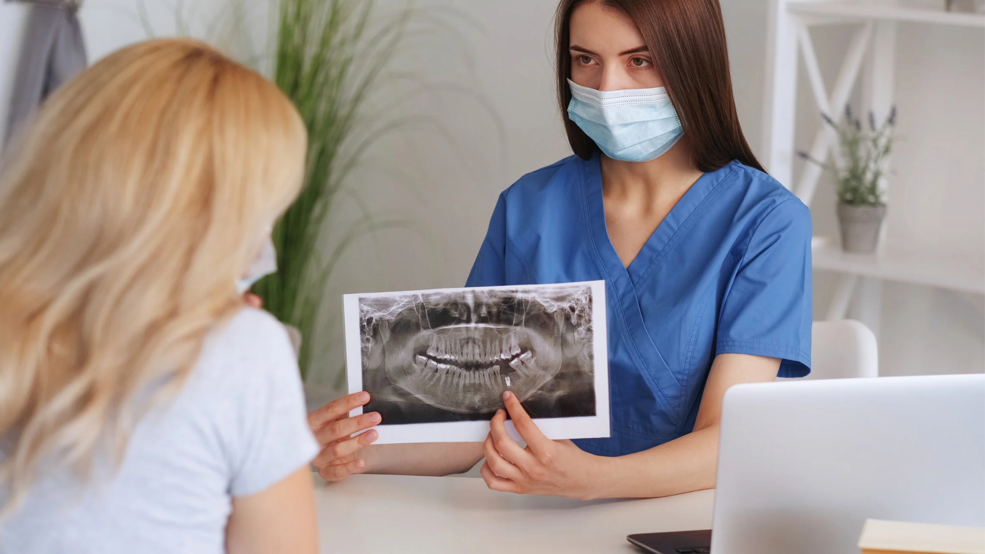 A masked dentist in blue scrubs shows a panoramic dental x-ray to a patient.