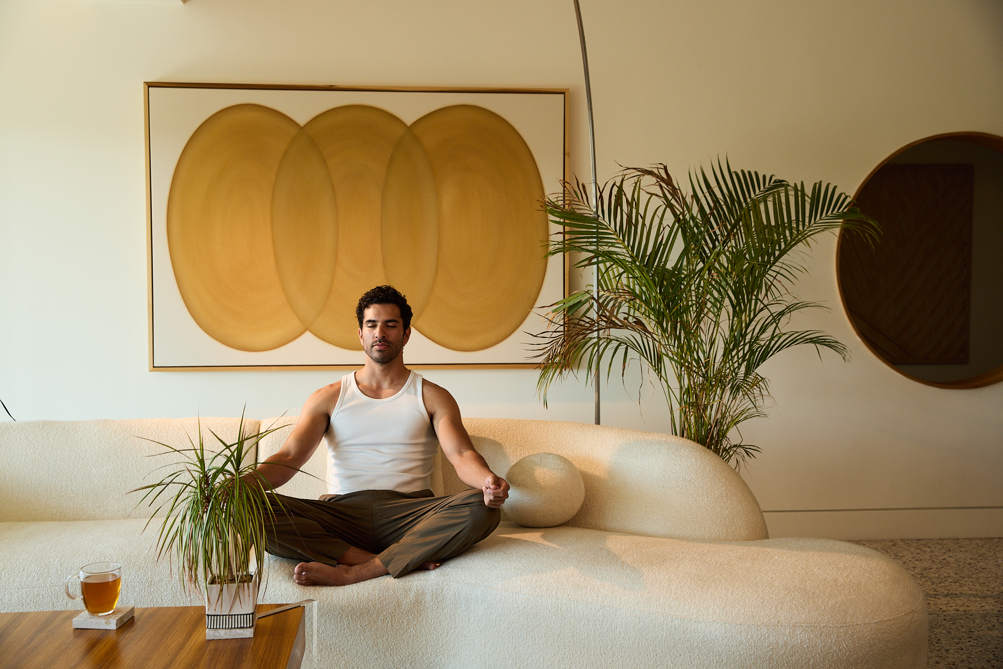 Person meditating on a sofa with plants and abstract art in the background.