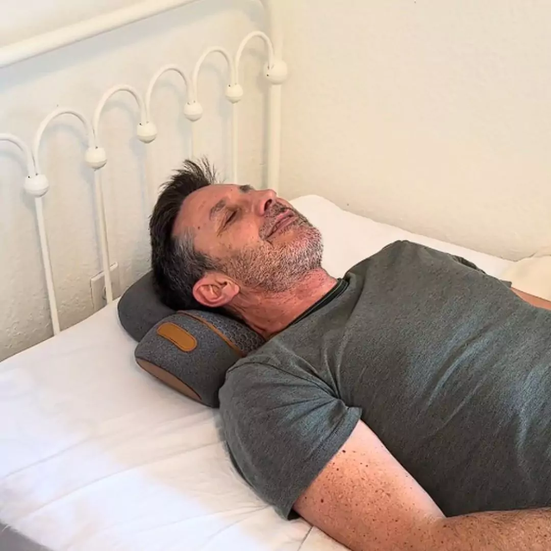 Man lying on a bed with a neck pillow, eyes closed.