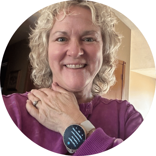 A smiling woman with curly blonde hair, wearing a purple sweatshirt and a smartwatch, looks at the camera.