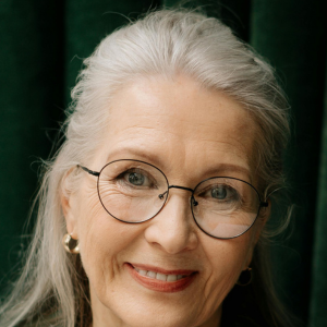 Elderly woman with glasses smiling against a dark green background.