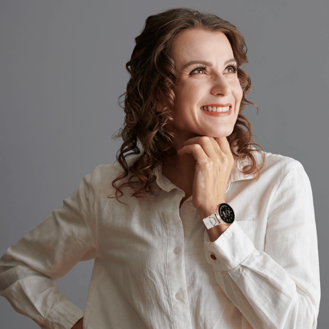 A smiling woman with curly brown hair in a white shirt looks up with her hand on her chin.