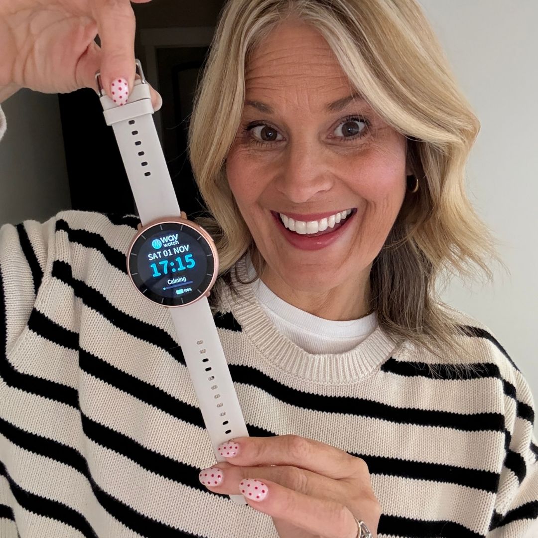 A smiling blonde woman in a striped sweater holds up a white smartwatch.