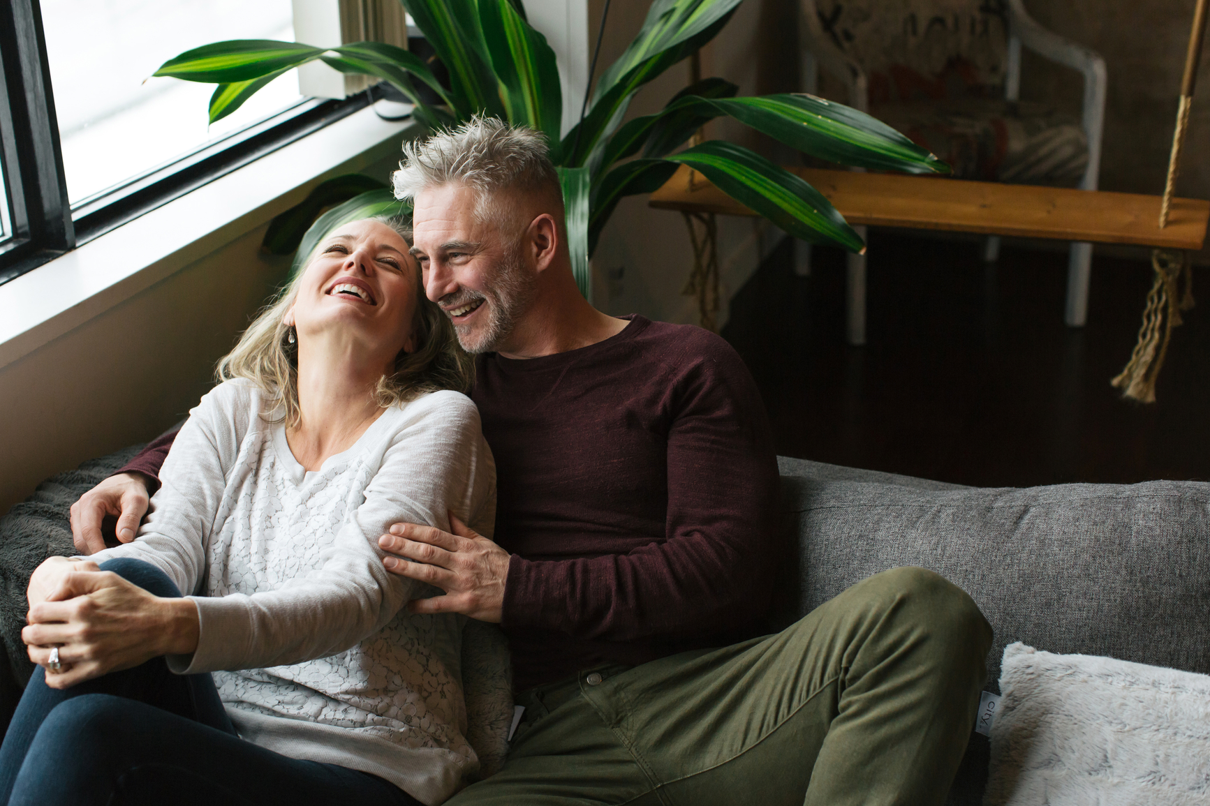 A smiling man with grey hair embraces a laughing woman as they sit together on a couch.