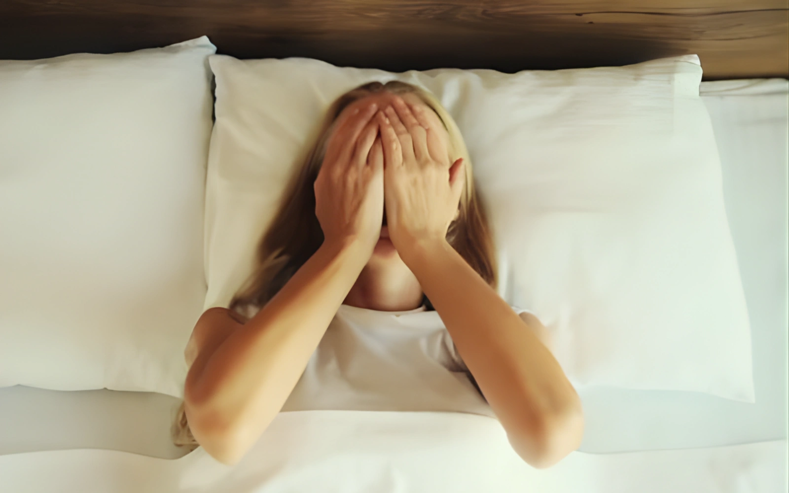 A person with long blonde hair lying in bed covers their face with their hands.