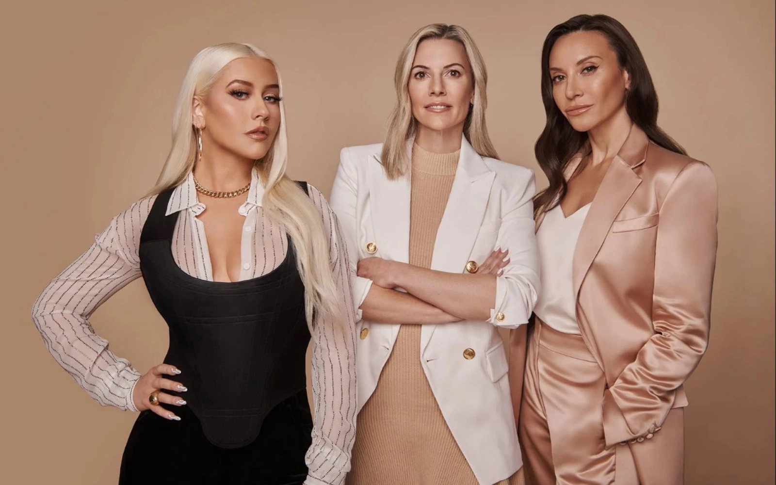 Three women in fashionable outfits pose together against a solid light brown background.