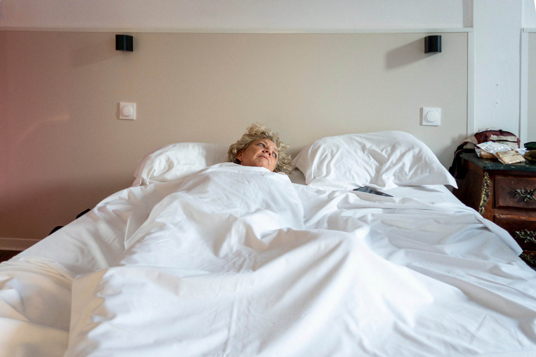 A woman with curly blonde hair lies in a bed with white linens, looking up and away.