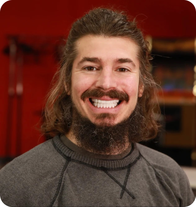Smiling man with long hair and beard, wearing a gray sweater, against a red background.