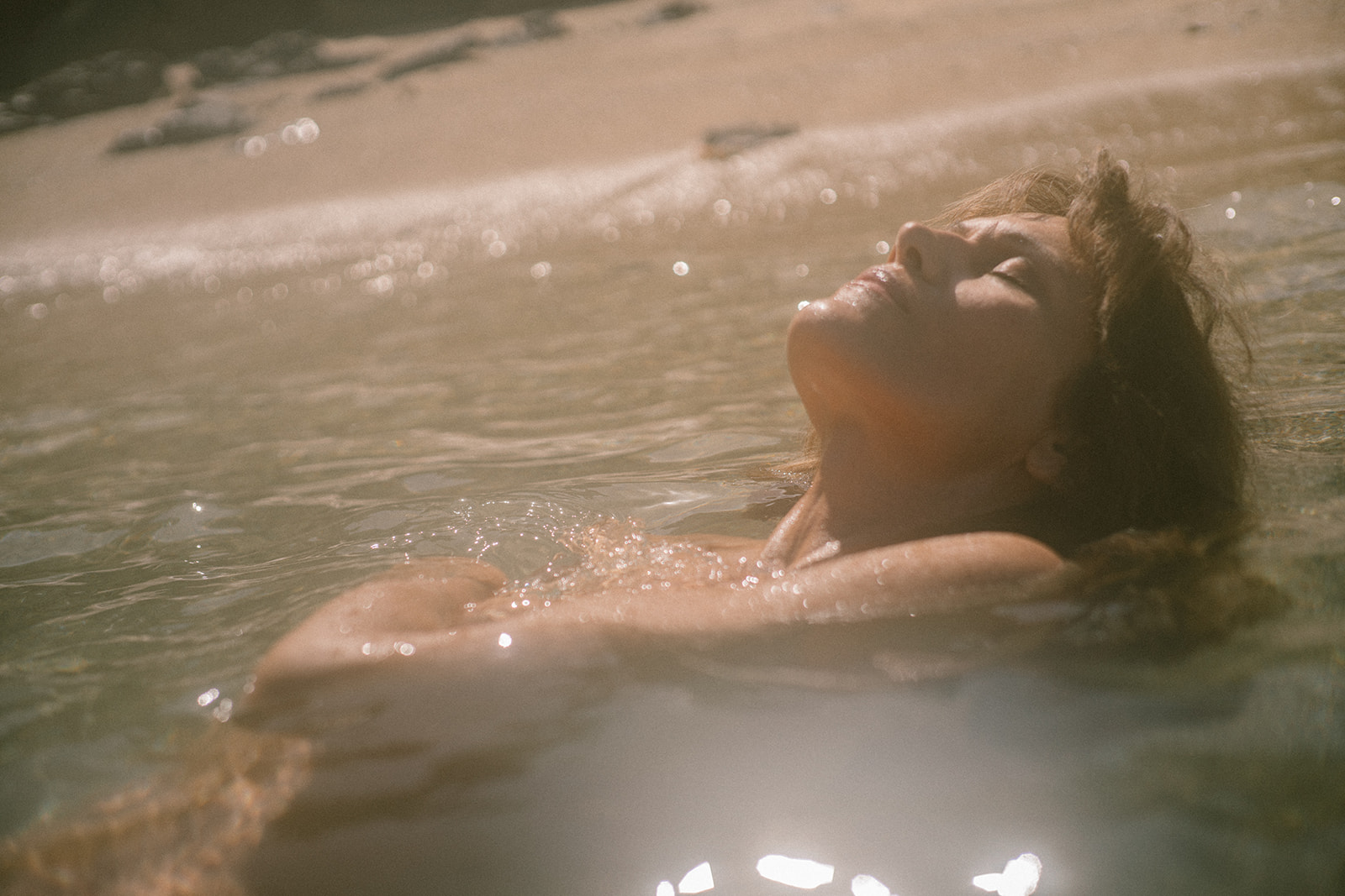 A woman with her eyes closed floats on her back in sun-dappled water near a sandy shore.