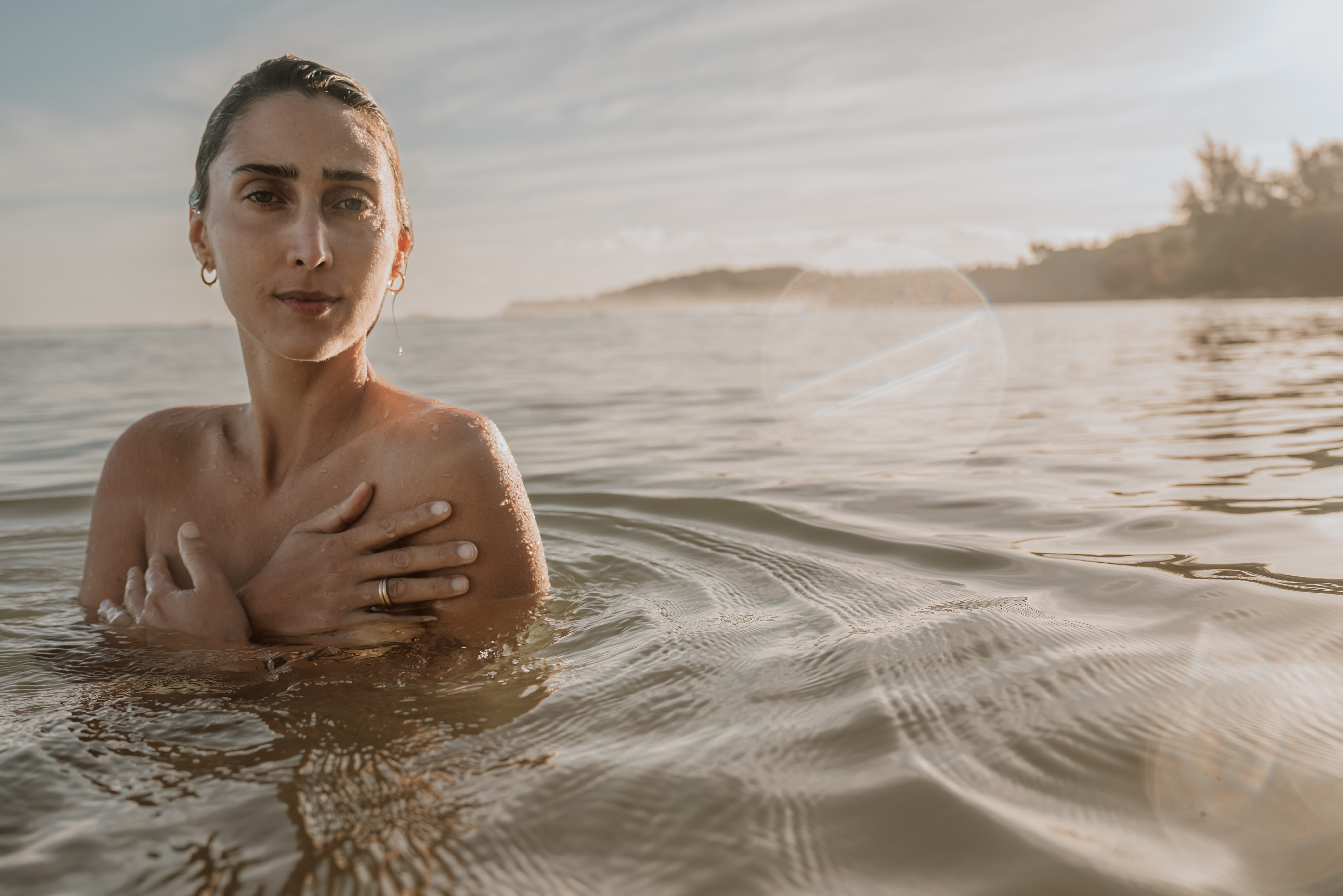 A woman stands chest-deep in the water at sunset, looking directly at the camera.