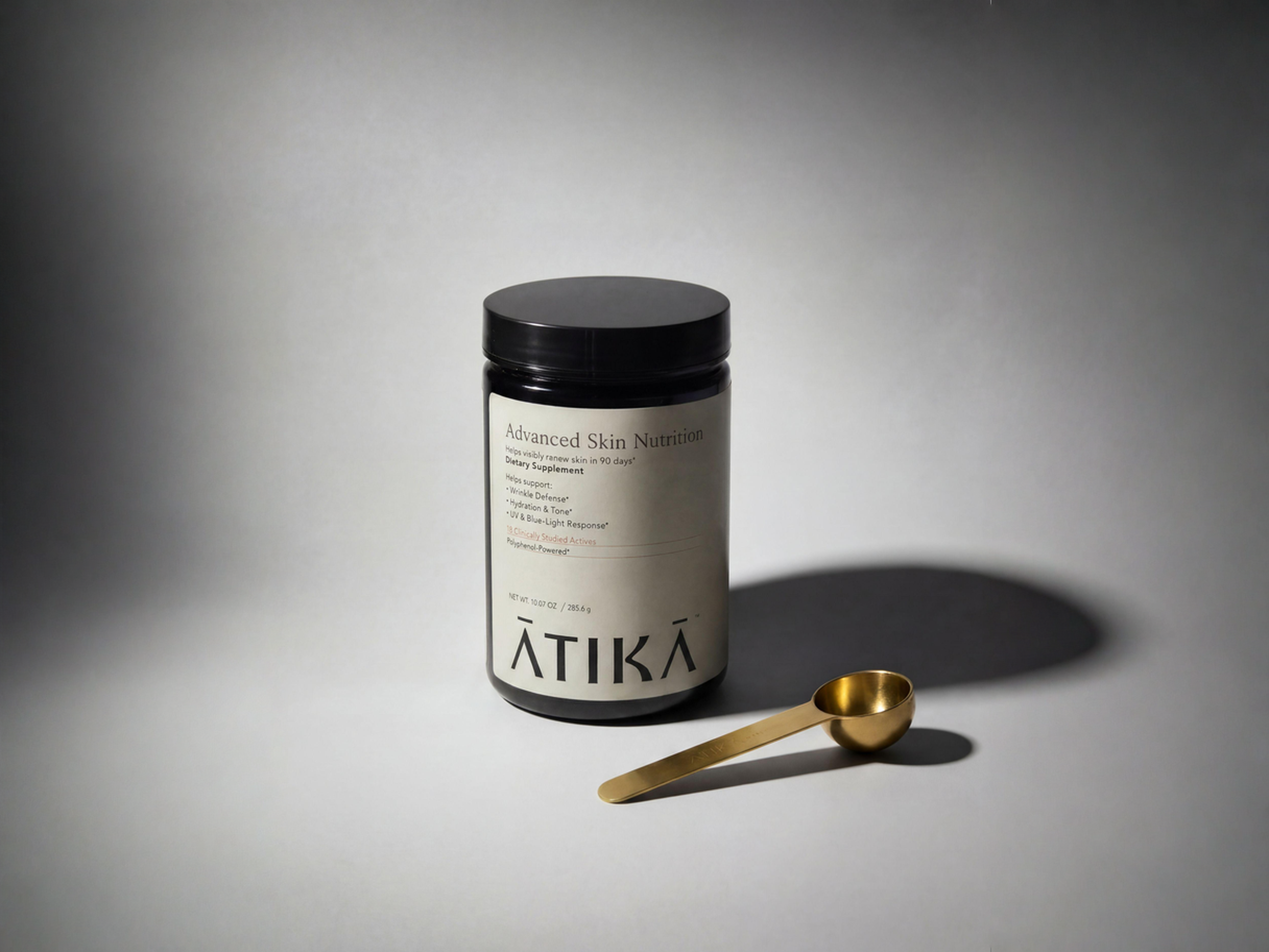 A black jar of Ātikā Advanced Skin Nutrition dietary supplement next to a small gold measuring spoon.