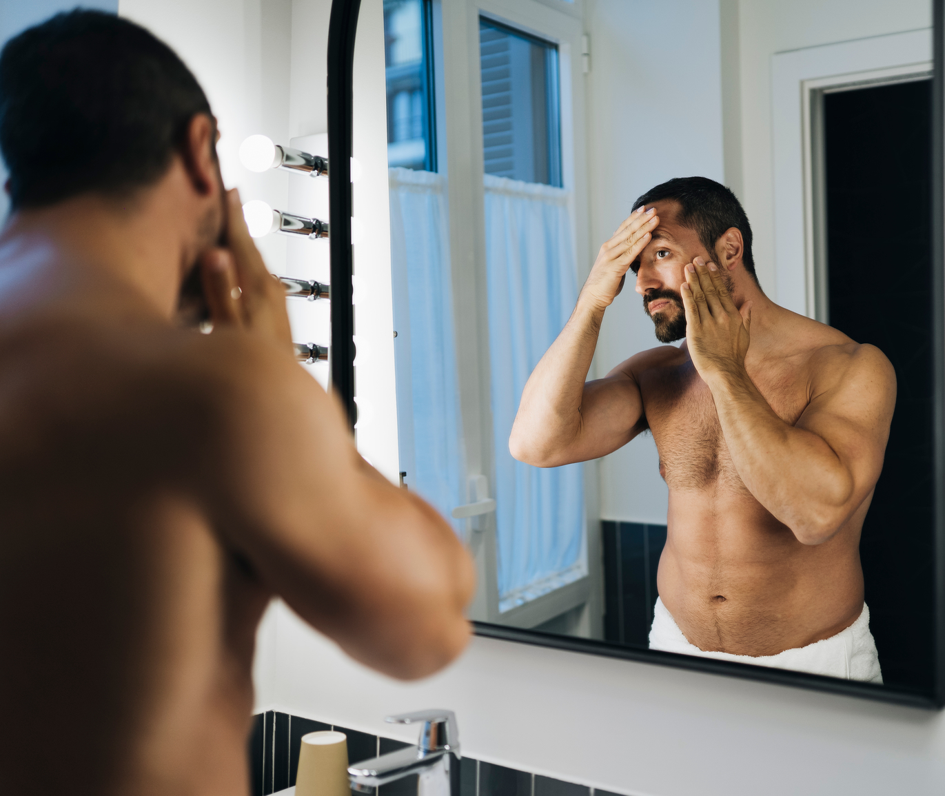 Man applying skincare in front of a bathroom mirror.