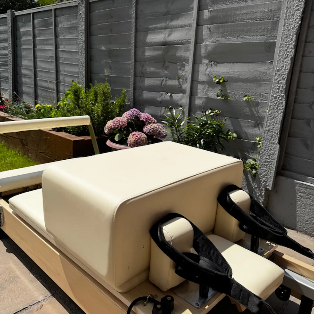Outdoor exercise equipment with padded seat and straps, near a wooden fence and garden.