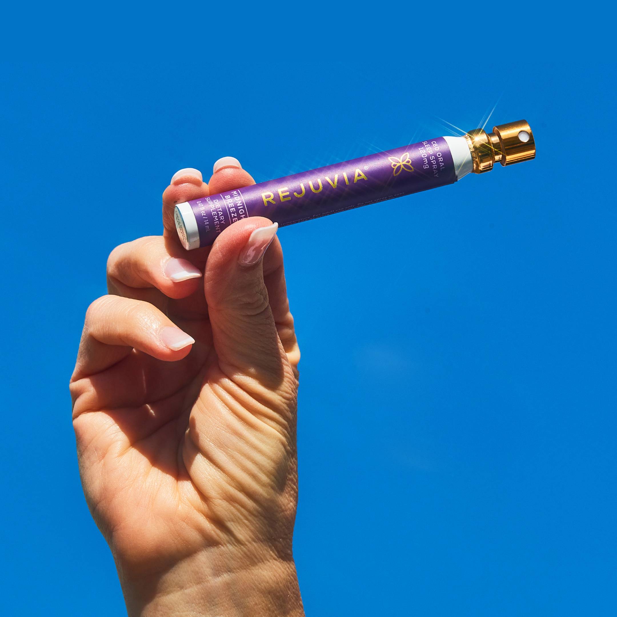 A hand holding a purple Rejuvia spray against a clear blue sky.