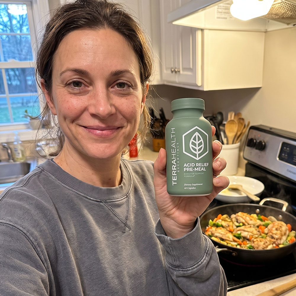 A smiling woman in a kitchen holds a bottle of Terrahealth Acid Relief Pre-Meal next to a stove.