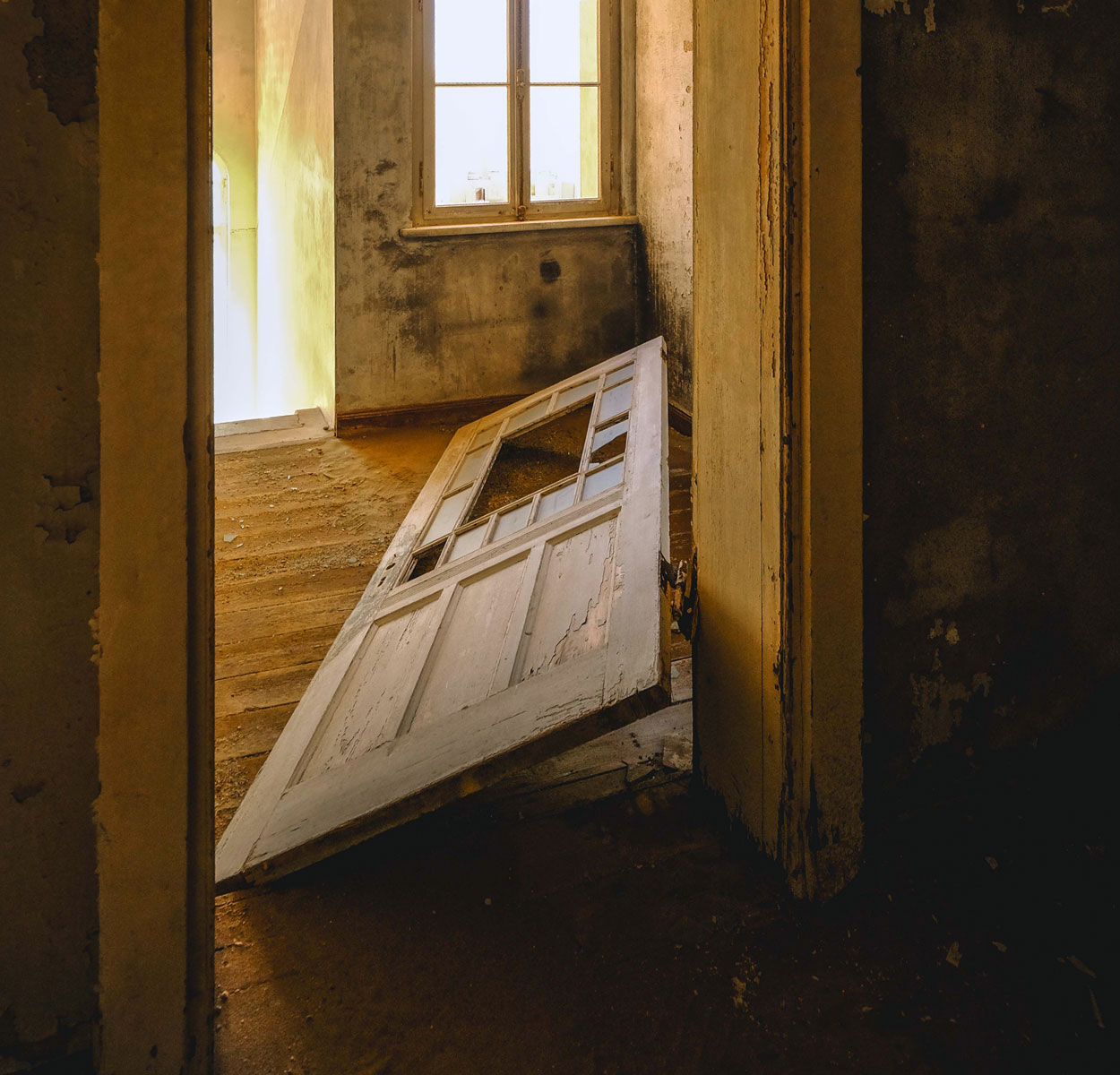 A white door with broken panes of glass lies on the floor of a dilapidated, sunlit room.