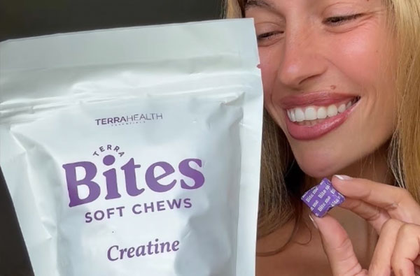 A smiling woman holds a purple creatine soft chew next to a bag of Terrahealth Bites.