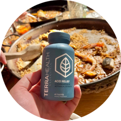 Hand holding a bottle labeled 'Terra Health Acid Relief' in front of a pan of food.
