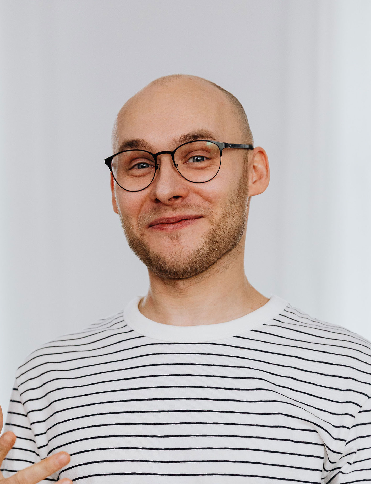 A bald man with glasses and a beard, wearing a striped shirt, smiling gently at the camera.