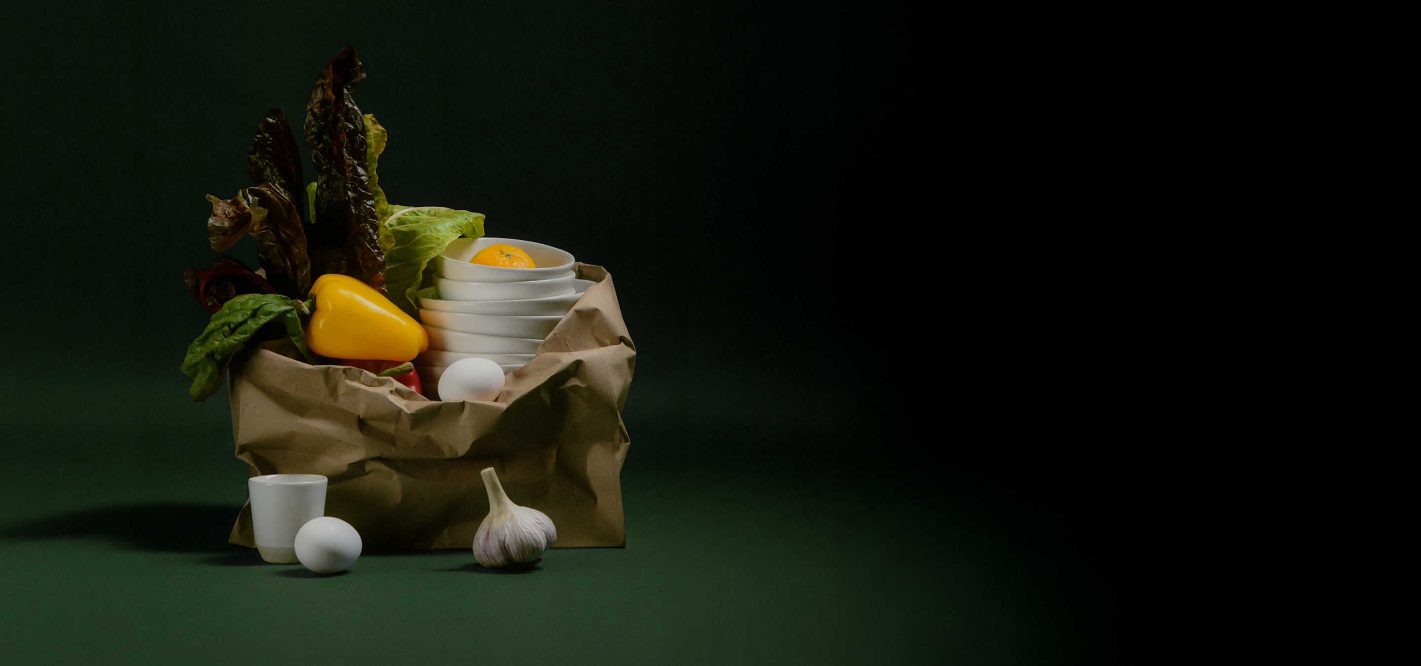Paper bag with vegetables, bowls, egg, and garlic on a dark background.