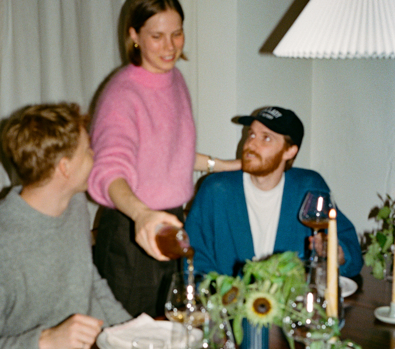 Three people at a table, one pouring wine, with glasses and sunflowers in view.