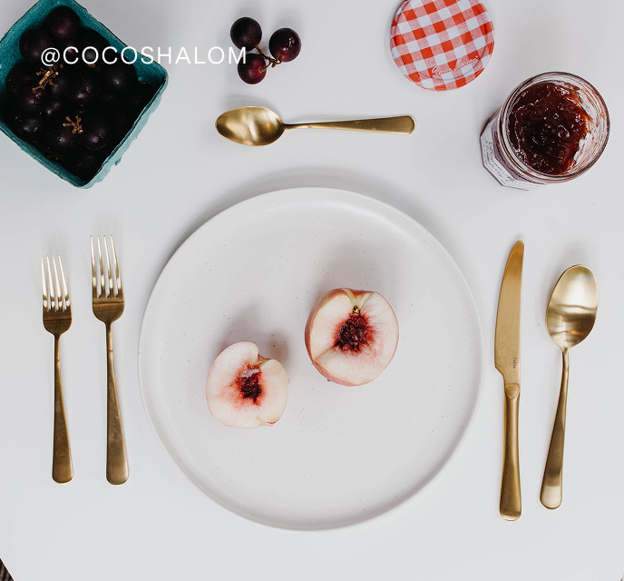 Styled flat lay with peaches, grapes, jam, and gold cutlery on a white table.