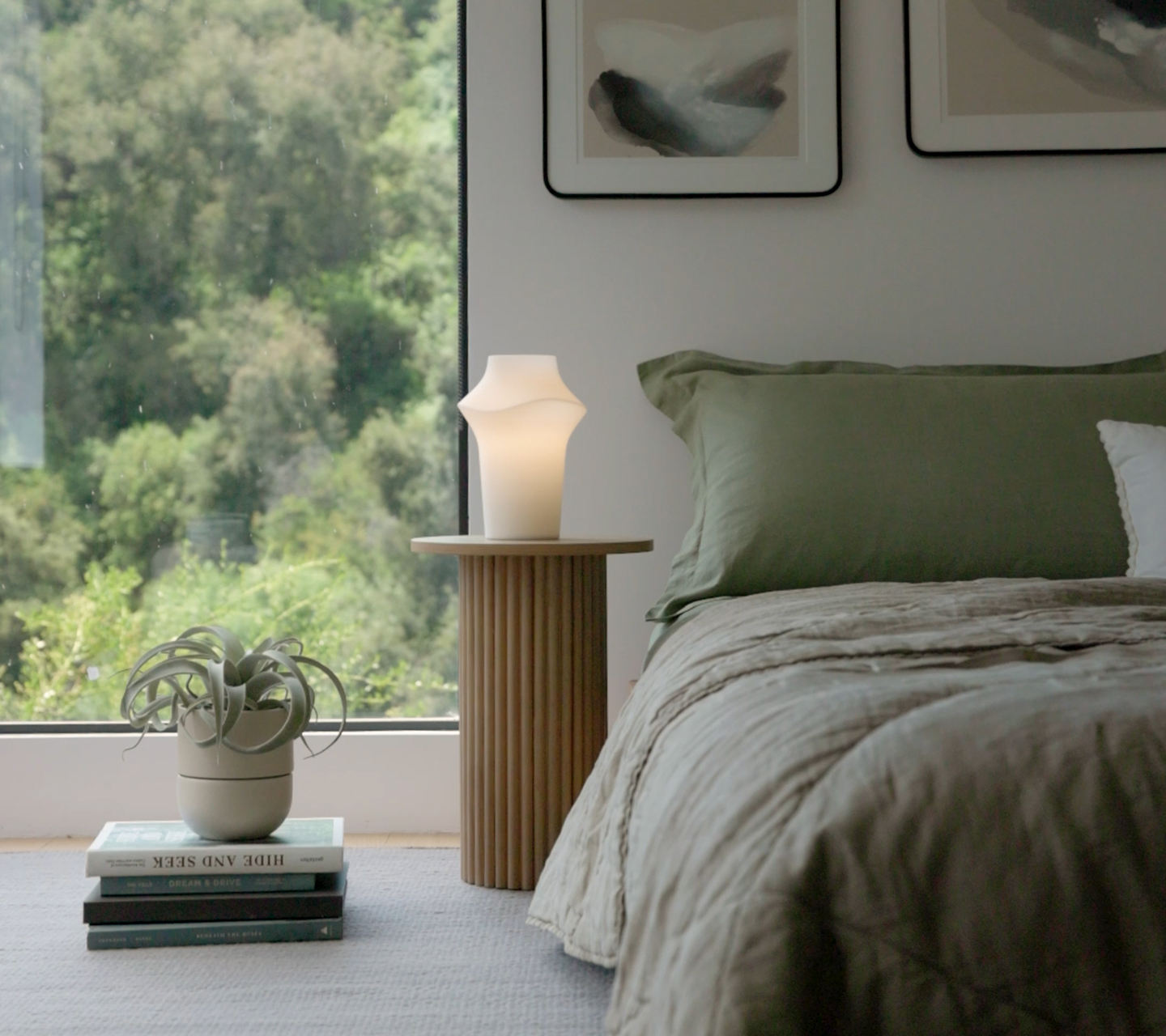 Bedroom with green bedding, a wooden nightstand, and a modern lamp by a large window.