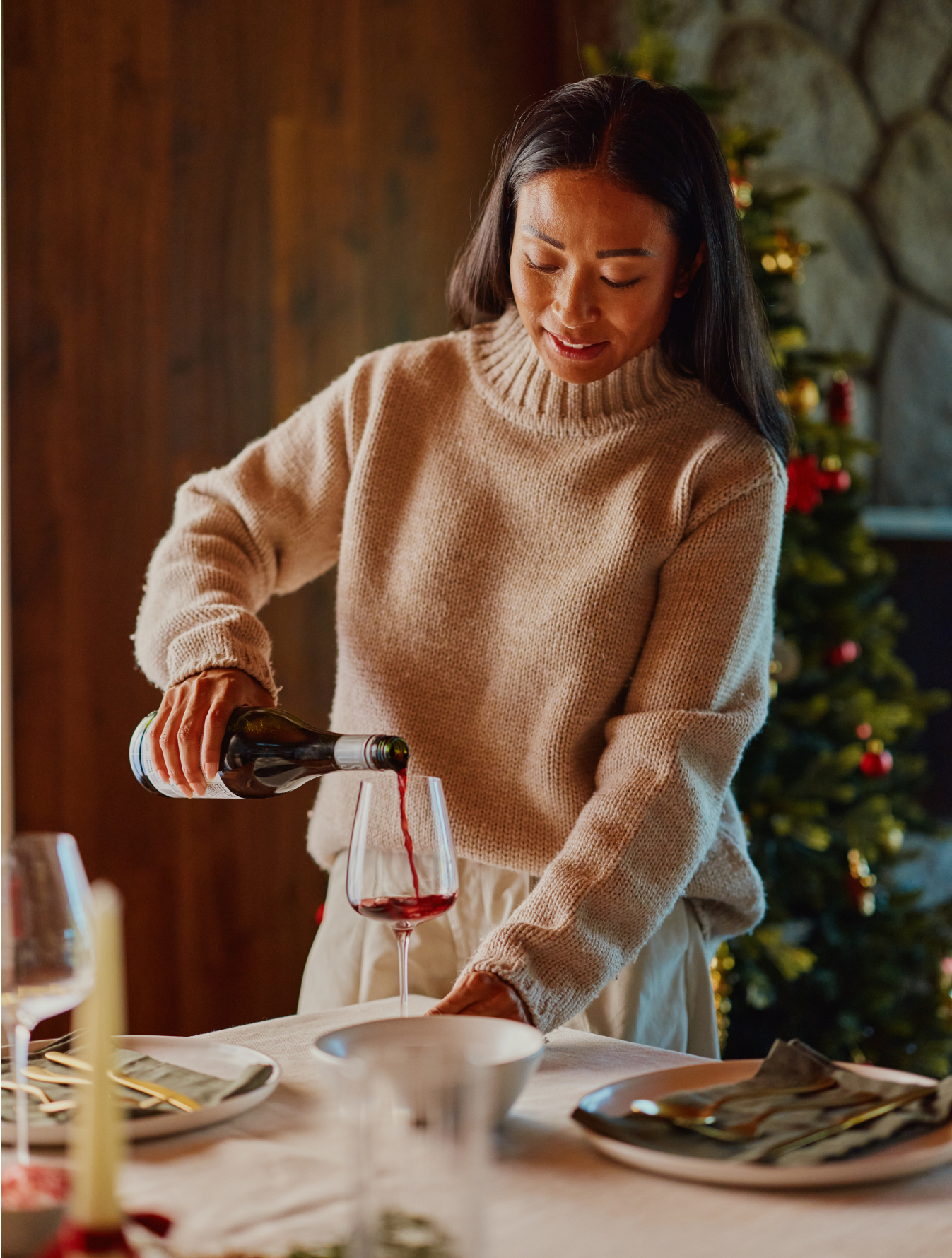 a woman standing at a festive dinner table, pouring red wine into a glass