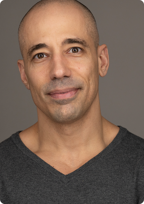 A person with a shaved head wearing a gray V-neck shirt, smiling slightly.