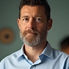 A person with a beard and mustache wearing a light blue shirt, standing indoors.