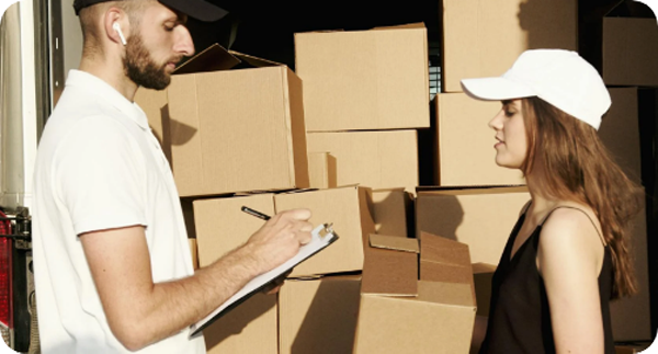 Two people with boxes, one holding a clipboard, outdoors.