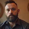 A person with a beard and short hair, wearing a dark shirt, in a softly lit room.