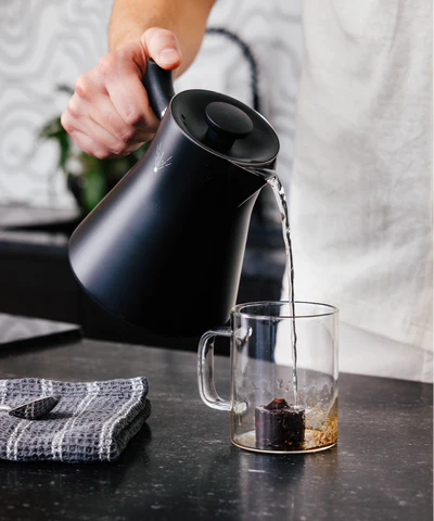 Person pouring hot water from a black kettle into a glass mug.