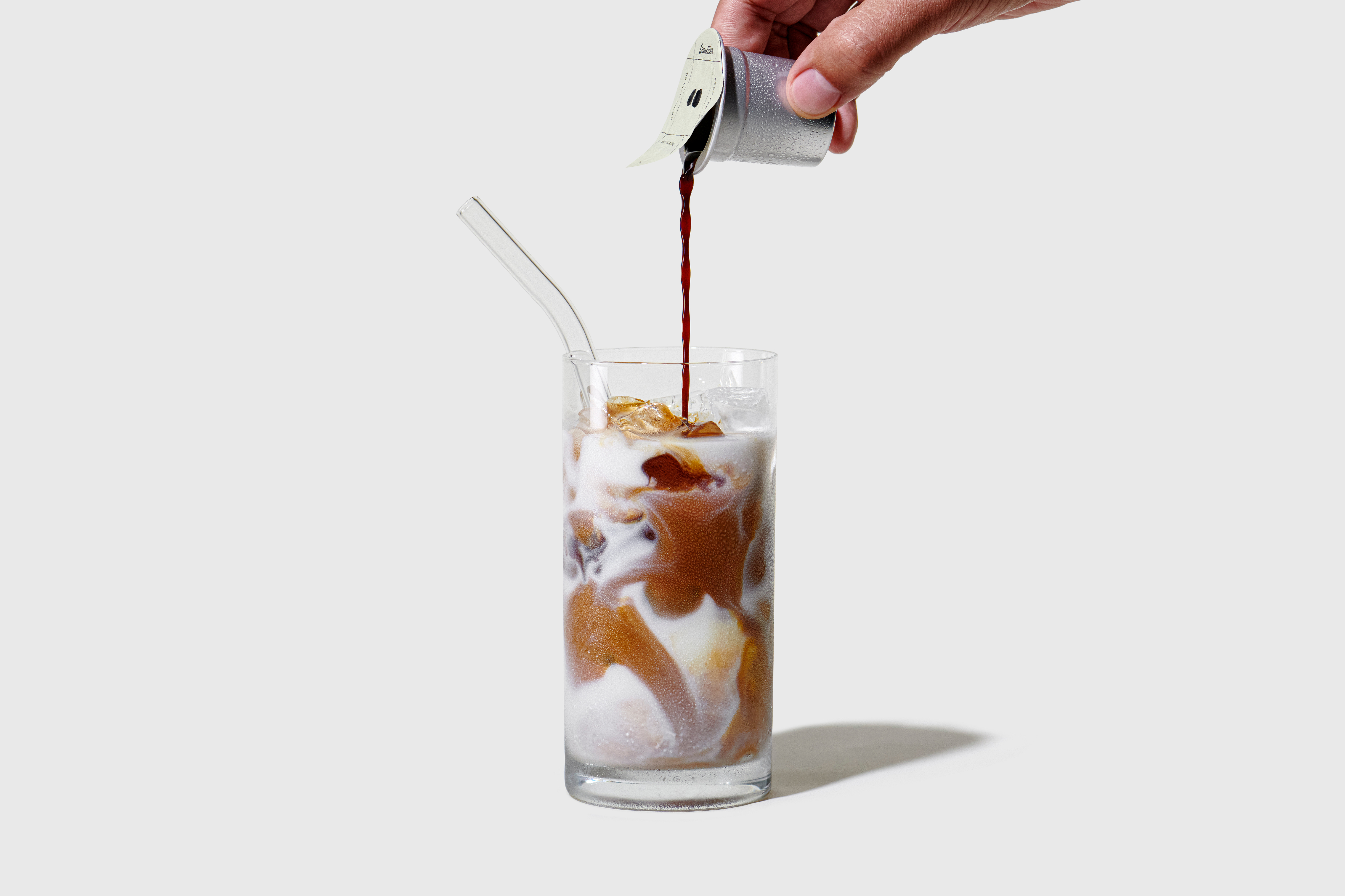 Hand pouring coffee into a glass of iced milk with a straw.