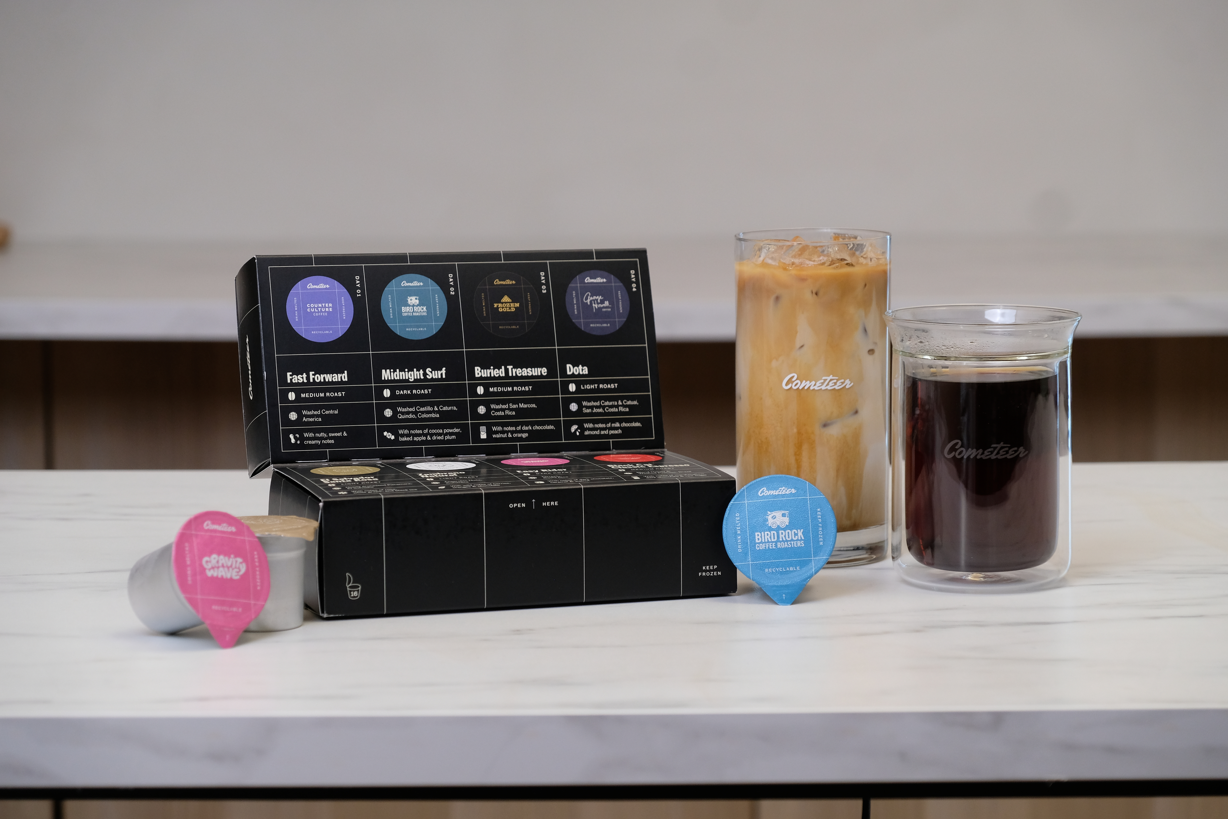 Coffee pods, iced coffee, and black coffee on a table with Cometeer branding.