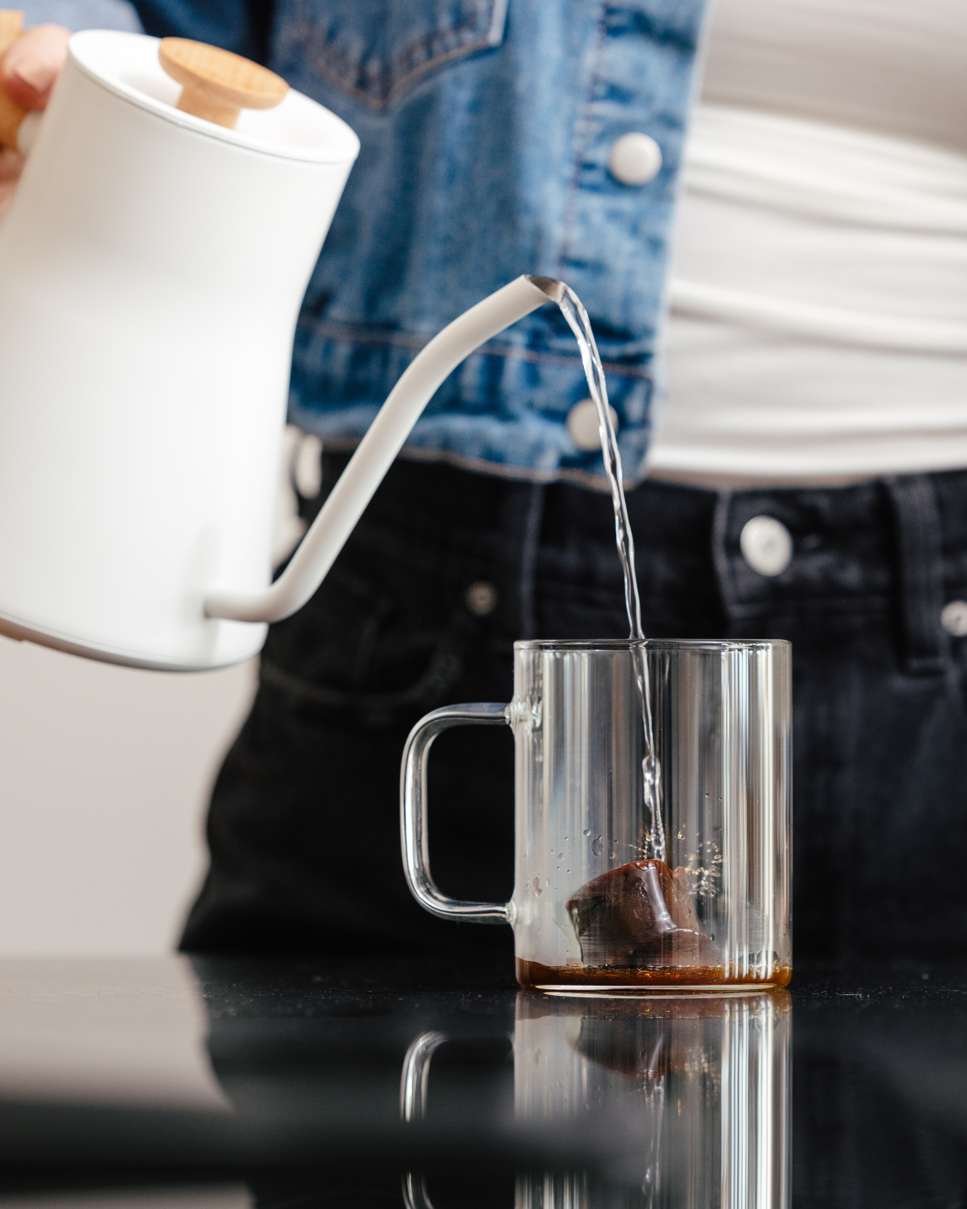 Person pouring water from a white kettle into a glass mug with a coffee cube.