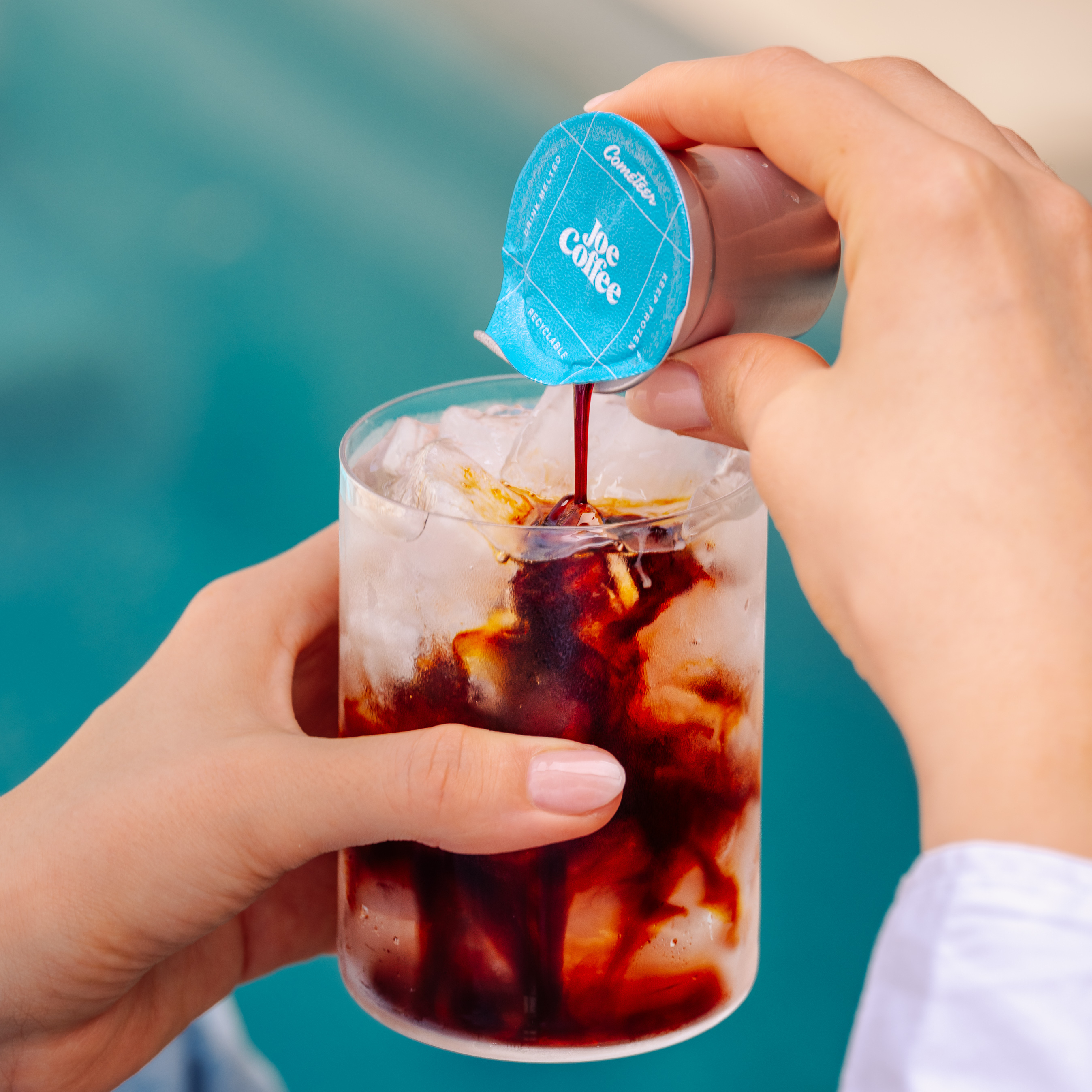 A person pours coffee concentrate from a small pod into a glass of ice.