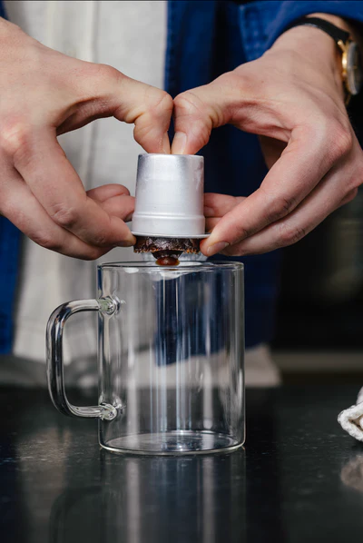 Hands pressing coffee capsule into a clear glass mug.